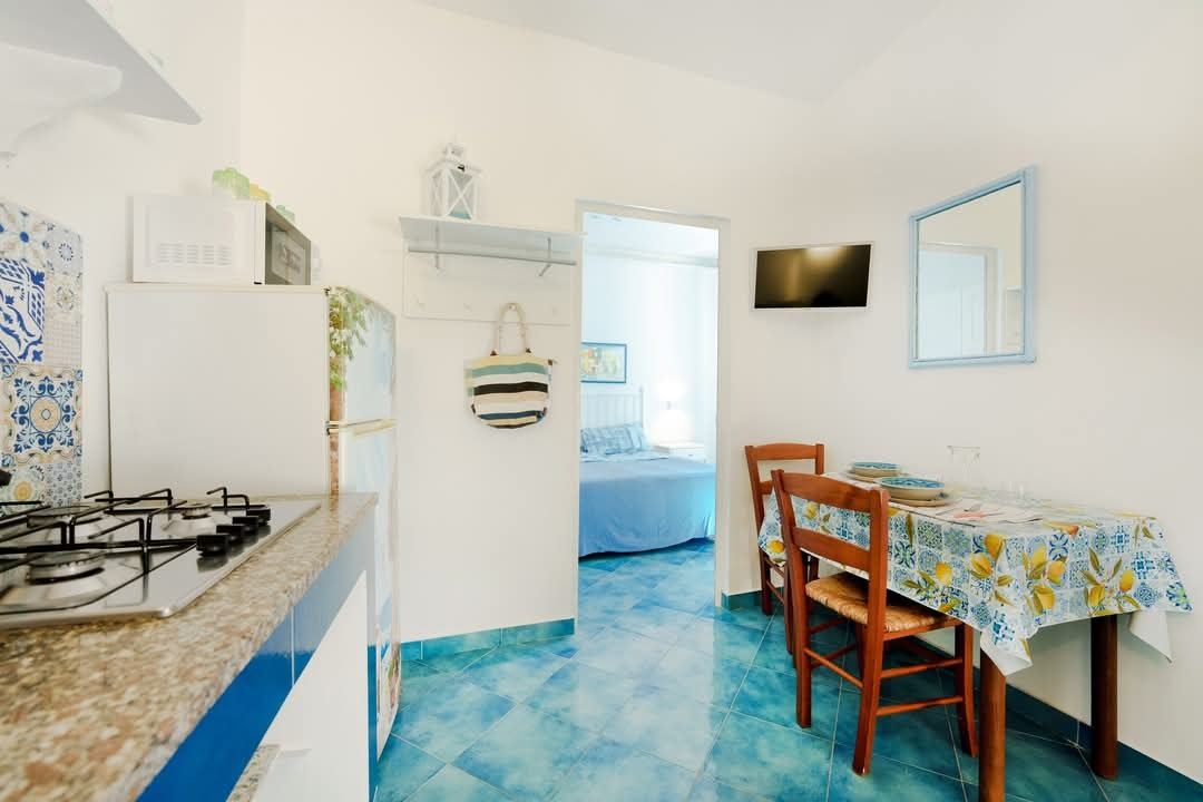 A bright, Mediterranean-style kitchenette and dining area with blue tile flooring, white appliances, and colorful patterned textiles.
