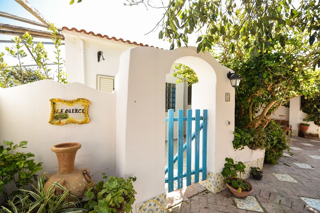 The charming whitewashed entrance gate with blue wooden door and decorative signage, surrounded by lush Mediterranean vegetation and terracotta accents.