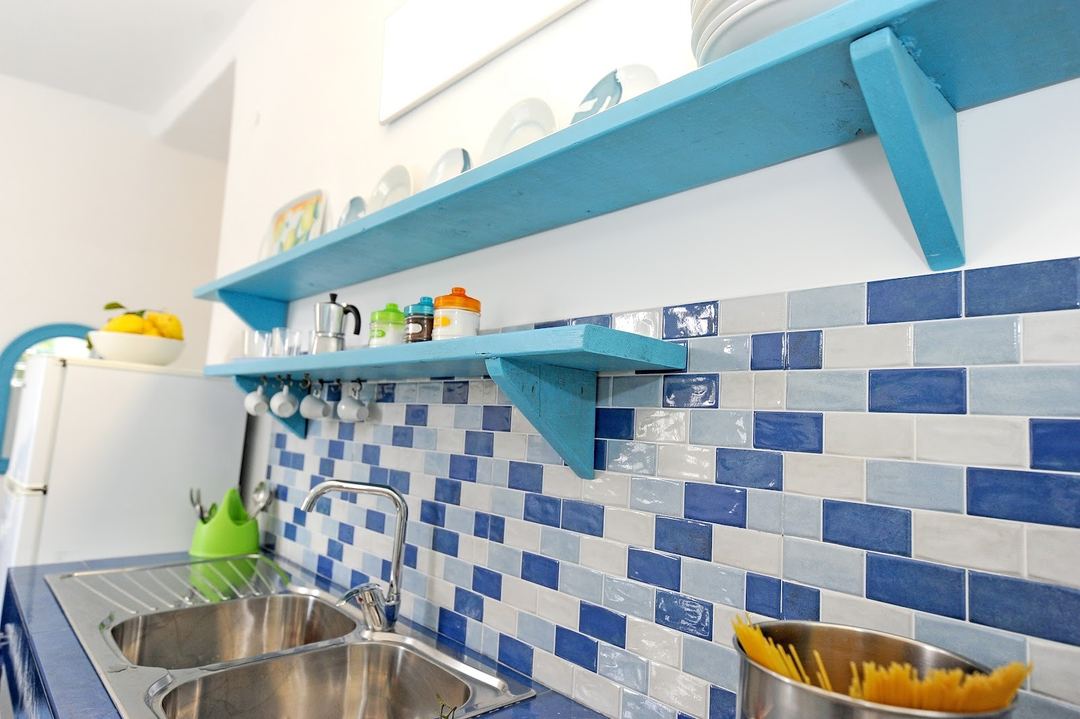 A bright kitchen detail featuring turquoise shelves with blue and white subway tile backsplash and stainless steel sink.