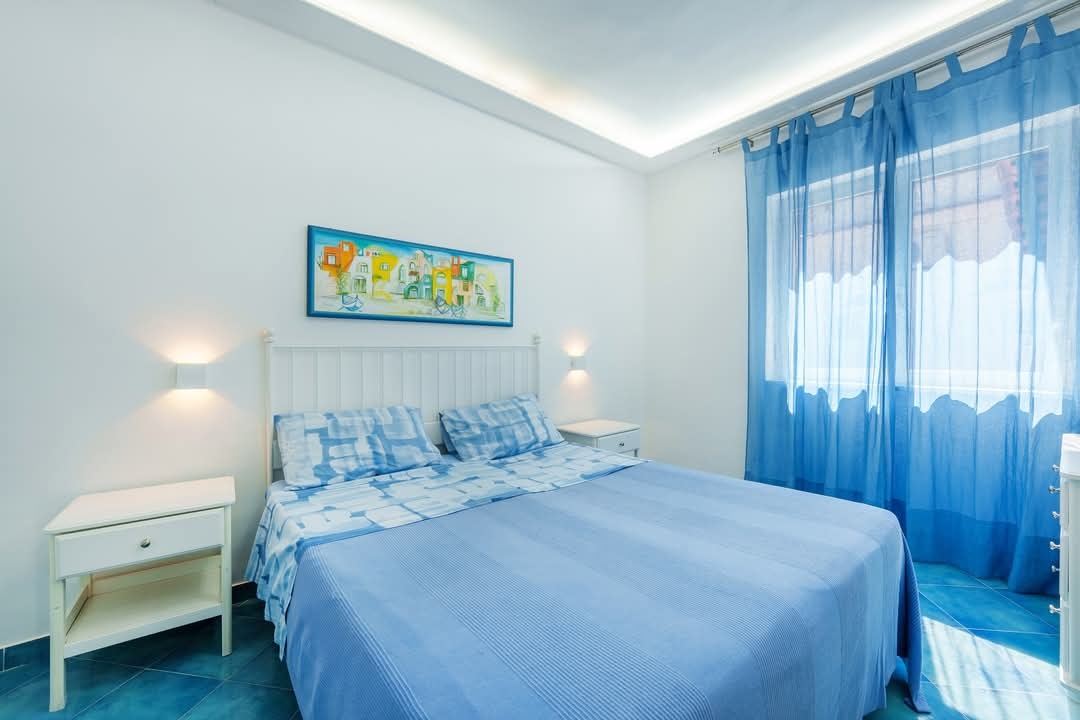 A bright, modern bedroom with blue and white bedding, white furniture, blue sheer curtains, and contemporary ceiling lighting.