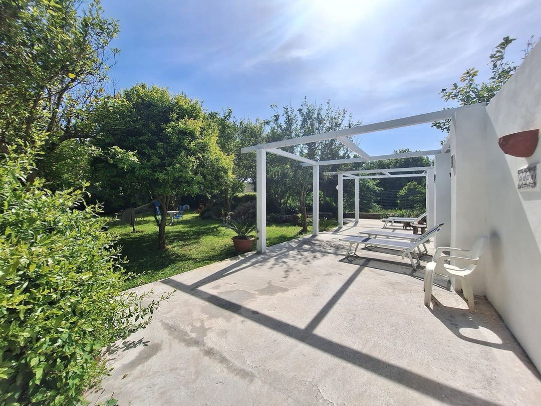 A modern white pergola with loungers overlooks the lush green gardens and trees surrounding the property.
