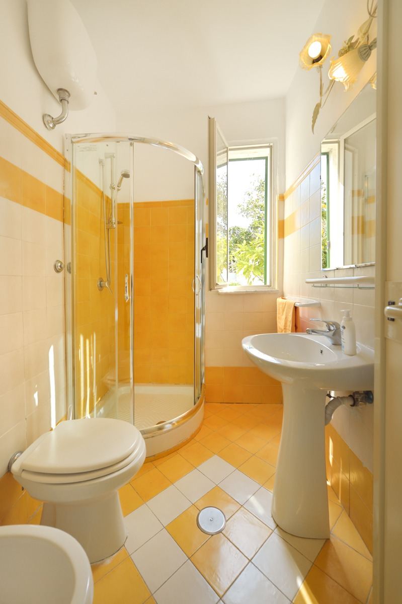 A bright bathroom featuring yellow and white tile work with a pedestal sink, toilet, and curved glass shower enclosure with a view to outdoor greenery.