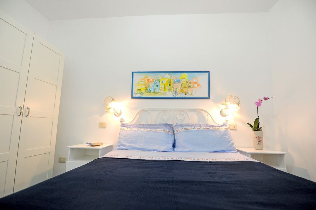 A bright, modern bedroom featuring a decorative wrought iron headboard with blue pillows, ambient wall sconces, and minimalist white furnishings.